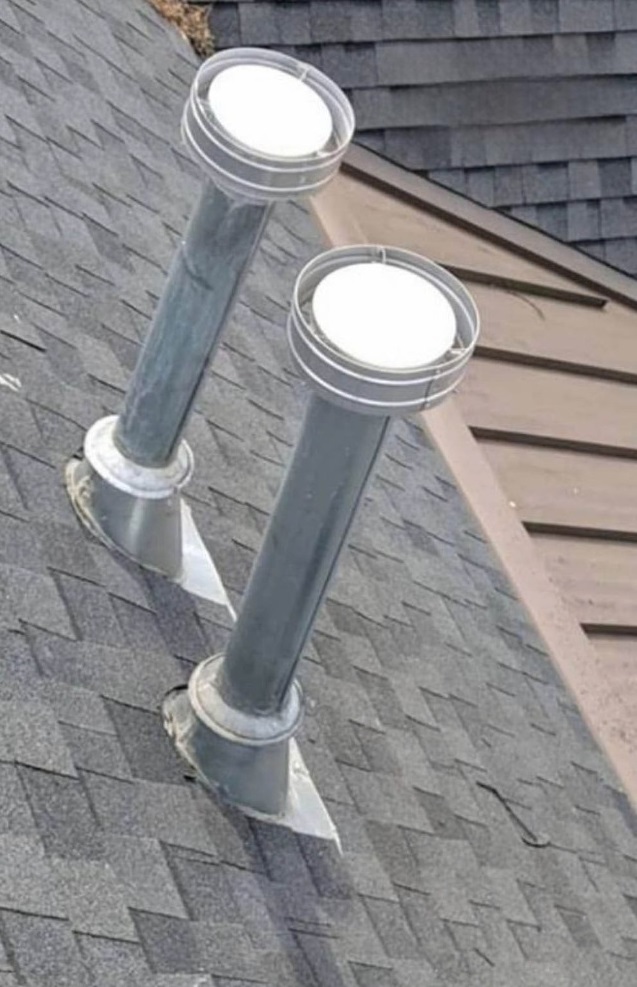 Two metal pipes are sticking out of a gray roof. They're like vents with round tops that let in light. The roof has gray shingles, and there's some brown wood visible too.