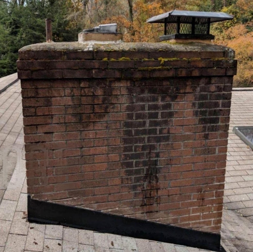 A brick chimney on a roof. It's made of red bricks, has a black metal thing on top (like a cap), and some moss is growing on it