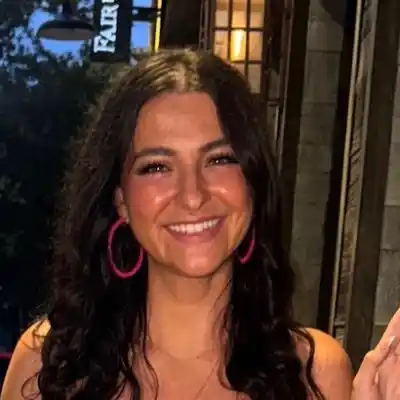 Abby Headshot A woman with long dark hair and pink hoop earrings is smiling in front of a building.