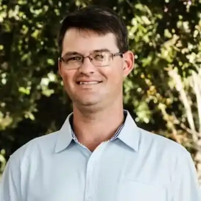 Rob Headshot The man is wearing a light blue shirt and glasses. He has short dark hair and is smiling. He is standing in front of trees.