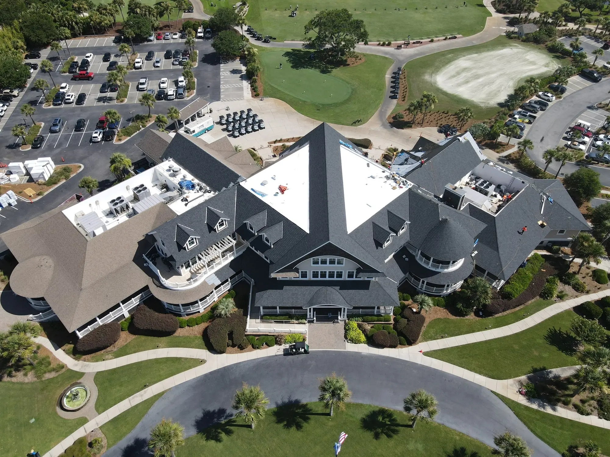 The building is a large clubhouse with a dark roof and white walls, surrounded by a golf course, parking lots, and landscaped areas with palm trees and grass.