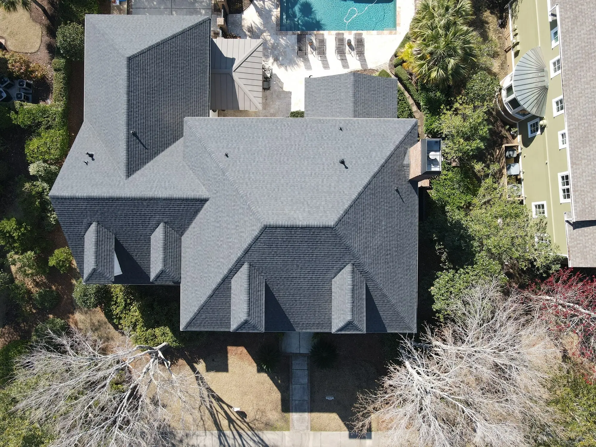 The house has a big gray roof and is surrounded by trees. There's a pool in the backyard with lounge chairs. Another house with green walls is nearby on the right.
