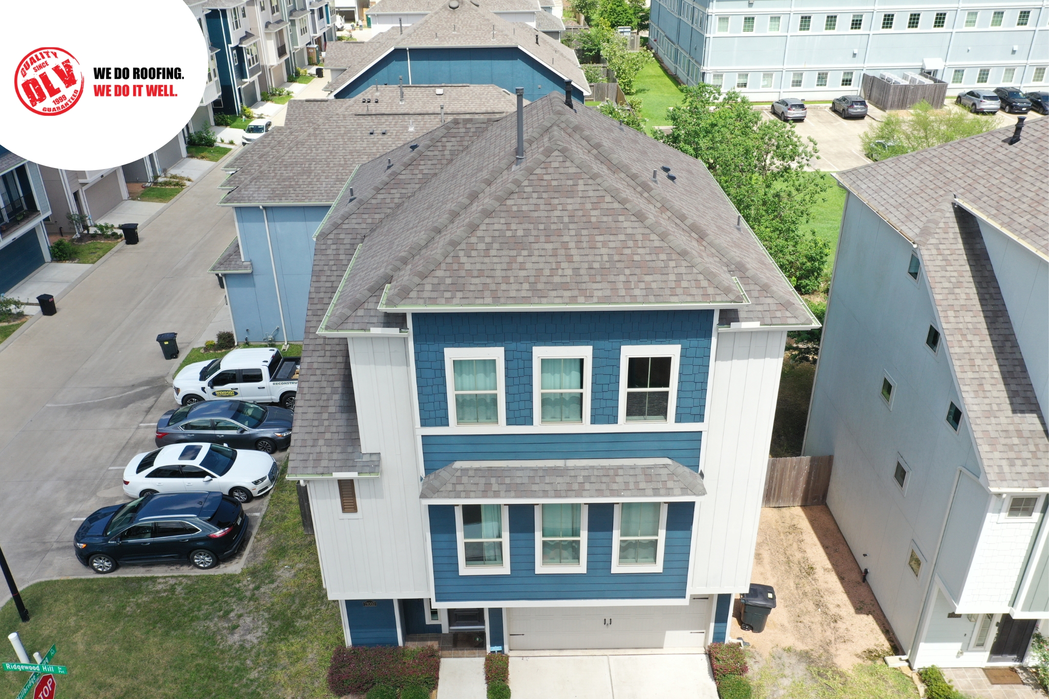 The house has a large front porch and is surrounded by trees and green grass. There's a driveway leading to the house, and a road is visible on the right side.